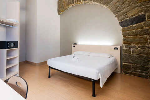 Minimalist hotel room at B&B Hotel Trieste with white bed linens, stone arch wall, wooden floor, and built-in shelving with a safe