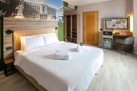 A modern hotel room at B&B Hotel Latina with a large bed with white linens, towels, wooden wardrobe, desk with chair, and TV on the wall