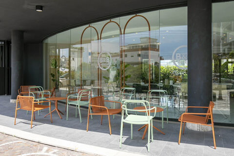Outdoor seating area with brown and light green metal chairs and tables outside B&B Hotel Latina with glass windows reflecting nearby buildings