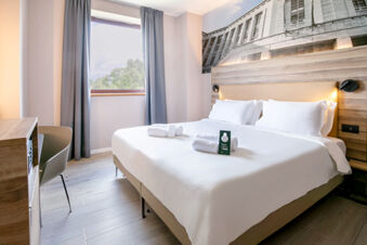 A clean hotel room at B&B Hotel Latina with a large bed, white linens, folded towels, wooden furniture, and a window with gray curtains