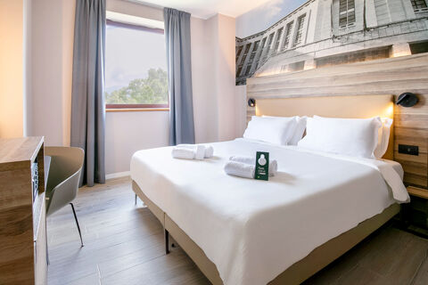 A clean hotel room at B&B Hotel Latina with a large bed, white linens, folded towels, wooden furniture, and a window with gray curtains