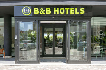 Entrance to B&B Hotel Latina with glass doors reflecting parked cars and outdoor seating visible inside the lobby area