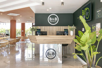 Reception desk with plants, two computers, and B&B Hotels logos at B&B Hotel Latina lobby with chairs and tables nearby