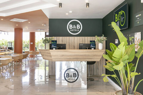 Reception desk with plants, two computers, and B&B Hotels logos at B&B Hotel Latina lobby with chairs and tables nearby