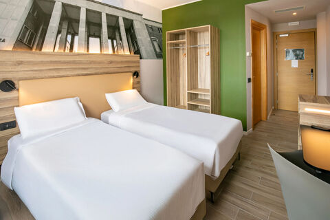 B&B Hotel Latina room with two single beds, wooden headboard, green accent wall, open closet, desk, and entrance door visible