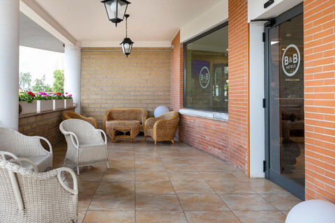 Outdoor seating area with wicker chairs, a small table, hanging lamps, flowers, and entrance door of B&B Hotel Pomezia Cortese