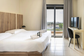 Hotel room at B&B Hotel Pomezia Cortese with two beds, white linens, bedside lamp, desk with TV, chair, and balcony door showing outside view