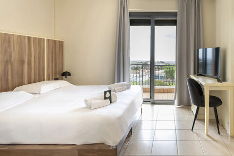Hotel room at B&B Hotel Pomezia Cortese with two beds, white linens, bedside lamp, desk with TV, chair, and balcony door showing outside view