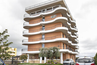 Exterior view of B&B Hotel Pomezia Cortese showing curved balconies, brick facade, parked cars, and surrounding trees and plants