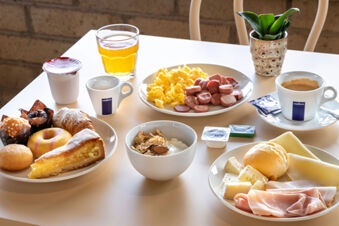 Breakfast spread on a table at B&B Hotel Pomezia Cortese featuring pastries, sliced sausages, scrambled eggs, cheese, cold cuts, yogurt, coffee, and juice
