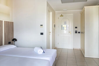 Minimalist hotel room interior at B&B Hotel Pomezia Cortese with a double bed, white linens, rolled towel, wardrobe, and closed door