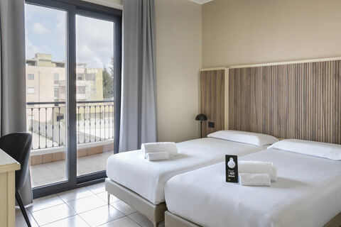 Two single beds with white linens and towels face a balcony with glass doors in the B&B Hotel Pomezia Cortese room
