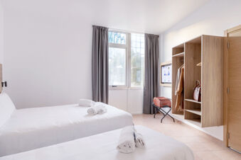 Two white beds with rolled towels face a window with gray curtains and wall-mounted cabinets in B&B Hotel Pomezia Roma