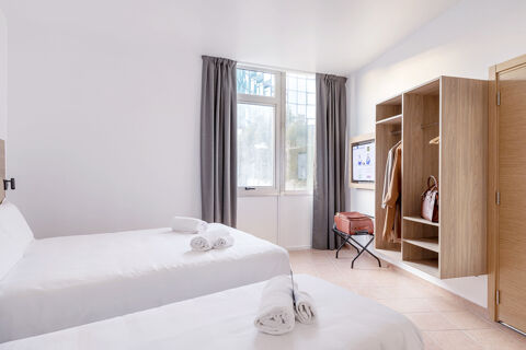Two white beds with rolled towels face a window with gray curtains and wall-mounted cabinets in B&B Hotel Pomezia Roma