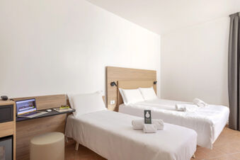 Two beds with white linens and towels in a bright room with desk, laptop and curtain at B&B Hotel Pomezia Roma