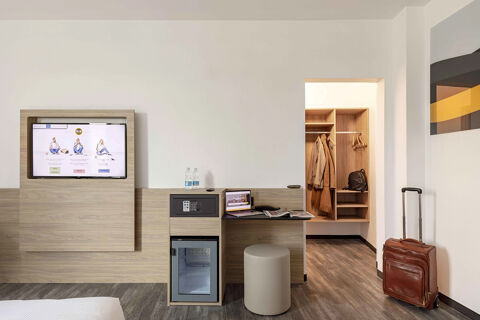 Hotel room interior at B&B Hotel Pomezia Roma with wall-mounted TV, desk, mini-fridge, open closet, and suitcase on floor