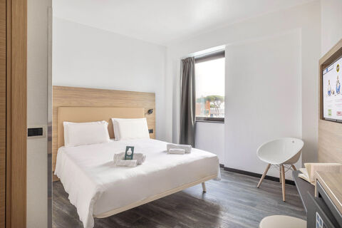 A modern hotel room at B&B Hotel Pomezia Roma with a double bed, white linens, a window, chair, TV, and wooden floor