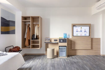Minimalist hotel room at B&B Hotel Pomezia Roma featuring open wooden shelving, wall-mounted TV, desk with laptop, and luggage rack