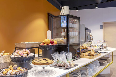 Breakfast buffet at B&B Hotel Pomezia Roma with fruit baskets, pastries, bread rolls, packaged cereals, and a refrigerated display case