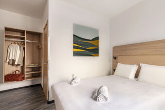 Minimalist hotel room at B&B Hotel Pomezia Roma with a double bed, white linens, rolled towels, and a small wardrobe with clothes and bag