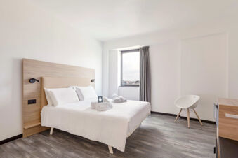 Bright hotel room at B&B Hotel Pomezia Roma with king bed, wooden headboard, rolled towels, window, and white chair