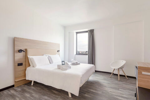 Bright hotel room at B&B Hotel Pomezia Roma with king bed, wooden headboard, rolled towels, window, and white chair