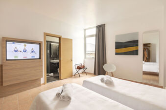 Bright hotel room at B&B Hotel Pomezia Roma with two beds, folded towels, a chair, and a bathroom visible through an open door
