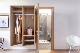 Open wooden wardrobe with hanging clothes and a brown handbag next to a bathroom door at B&B Hotel Pomezia Roma