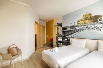 Modern hotel room with two white beds, a chair with a bag, open wooden door, and a mural at B&B Hotel Roma Fiumicino Aeroporto Fiera 2