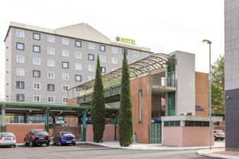 B&B Hotel Roma Fiumicino Aeroporto Fiera 2 building with multiple windows, two tall cypress trees, and parked cars nearby