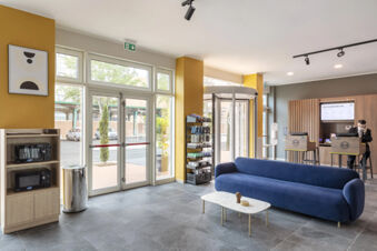 Lobby area of B&B Hotel Roma Fiumicino Aeroporto Fiera 2 showing blue sofa, reception desks, large windows, and microwave shelves