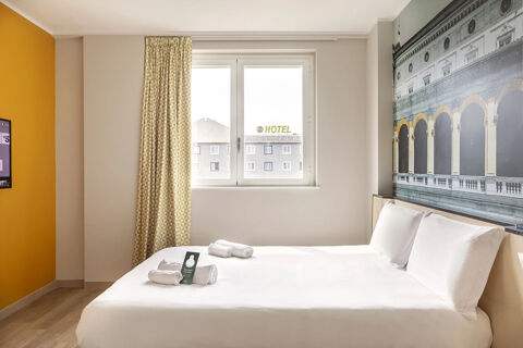 Bedroom with white bedding, yellow patterned curtains, a large window, wall art, and towels at B&B Hotel Roma Fiumicino Aeroporto Fiera 2