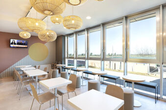 Dining area with white tables, wooden chairs, round stools, large windows, and hanging wicker light fixtures at B&B Hotel Roma Fiumicino Aeroporto Fiera