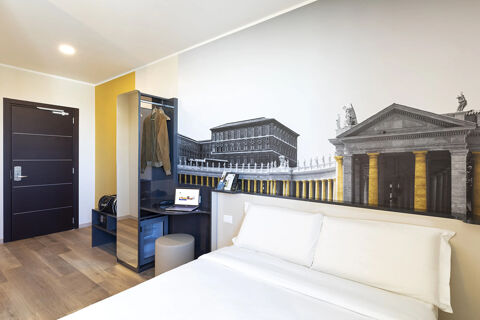 Hotel room at B&B Hotel Roma Fiumicino Aeroporto Fiera with white bed, dark wood door, mural of classical building, and small desk with laptop