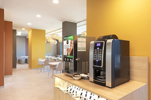 Two beverage vending machines on a wooden counter with glasses and cups in the B&B Hotel Roma Fiumicino Aeroporto Fiera dining area