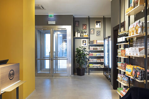 Interior view of B&B Hotel Roma Fiumicino Aeroporto Fiera lobby with yellow wall, glass doors, snack shelves, and framed pictures on wall