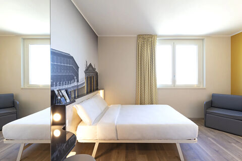 Bright hotel room at B&B Hotel Roma Fiumicino Aeroporto Fiera with double bed, gray sofas, patterned curtains, and wall art