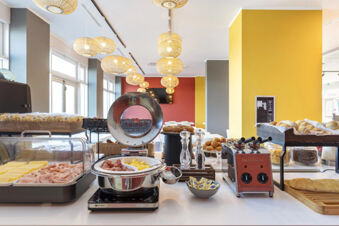 Breakfast buffet at B&B Hotel Roma Fiumicino Aeroporto Fiera with sliced ham, cheese, scrambled eggs, rolls, and kitchen appliances visible