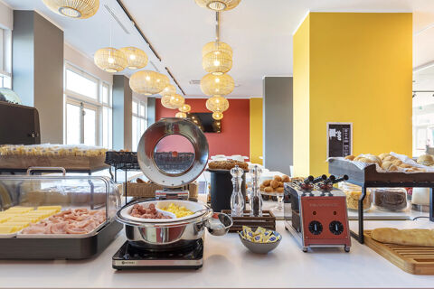 Breakfast buffet at B&B Hotel Roma Fiumicino Aeroporto Fiera with sliced ham, cheese, scrambled eggs, rolls, and kitchen appliances visible