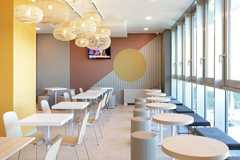 Bright dining area with white tables and chairs, gray ottomans, large windows, and hanging rattan lamps at B&B Hotel Roma Fiumicino Aeroporto Fiera