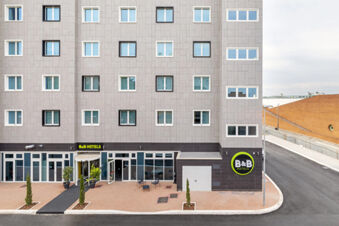 Facade of B&B Hotel Roma Fiumicino Aeroporto Fiera with multiple windows, black and white doors, small trees, and outdoor seating