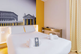 Modern hotel room with a large white bed, folded towels, yellow accents, a black and white mural, and a curtain at B&B Hotel Roma Pietralata Tiburtina