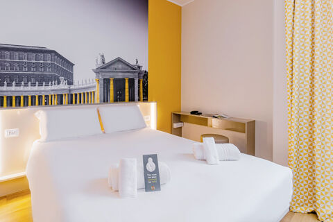 Modern hotel room with a large white bed, folded towels, yellow accents, a black and white mural, and a curtain at B&B Hotel Roma Pietralata Tiburtina