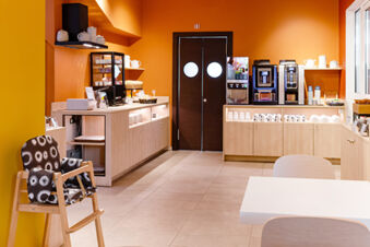Small dining area with a high chair, coffee machines, cups, plates, and snack display at B&B Hotel Roma Pietralata Tiburtina