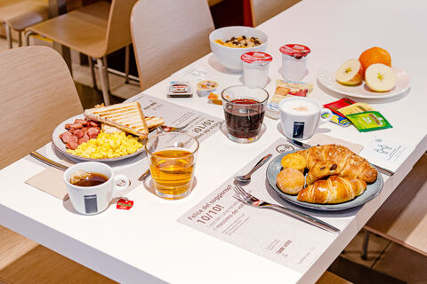 Breakfast spread on a white table at B&B Hotel Roma Pietralata Tiburtina with scrambled eggs, toast, croissants, tea, coffee, and fruit