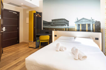 Hotel room at B&B Hotel Roma San Lorenzo Termini with a double bed, rolled towels, yellow chair, and wall mural of historic buildings