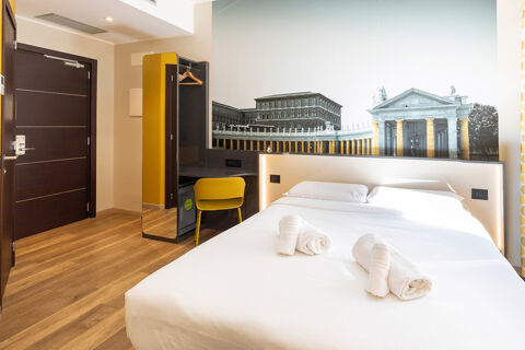 Hotel room at B&B Hotel Roma San Lorenzo Termini with a double bed, rolled towels, yellow chair, and wall mural of historic buildings
