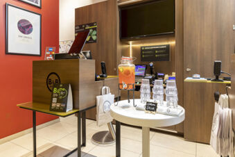 Reception desk with a beverage dispenser, stacked plastic cups, and framed maps at B&B Hotel Roma San Lorenzo Termini