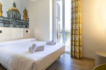 Bedroom with a double bed with white sheets and towels, a wall mural of a building, and yellow patterned curtains at B&B Hotel Roma San Lorenzo Termini