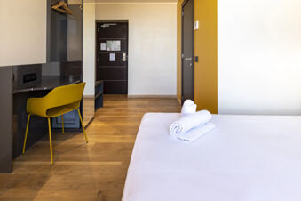 A modern hotel room at B&B Hotel Roma San Lorenzo Termini with a white bed, folded towels, yellow chair, and wooden floor